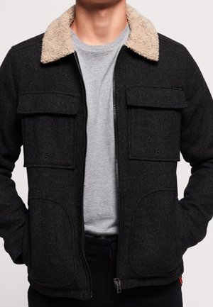 Mand iført en mørkegrå jakke med lynlås, beige sherpa-krave og store forlommer over en lysegrå t-shirt.