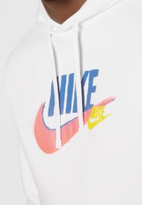 Närbild av vit huvtröja med blåa, rosa och gula Nike-logotyper på bröstet och vita dragsnören.