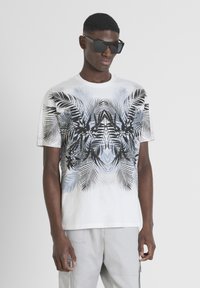 Camiseta blanca de manga corta con un patrón de hojas de palma en negro y azul, combinada con gafas de sol negras angulares. Textura de algodón suave.