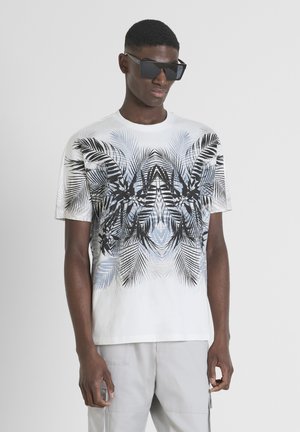 Camiseta blanca de manga corta con un patrón de hojas de palma en negro y azul, combinada con gafas de sol negras angulares. Textura de algodón suave.