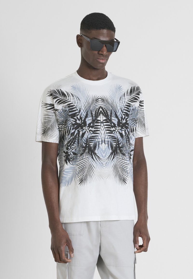 Camiseta blanca de manga corta con un patrón de hojas de palma en negro y azul, combinada con gafas de sol negras angulares. Textura de algodón suave.