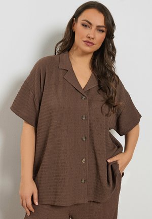 Frau mit langen dunklen Haaren, die ein lockeres, strukturiertes braunes kurzärmeliges Hemd mit Knöpfen und passende Hose trägt, stehend mit einer Hand in der Tasche.