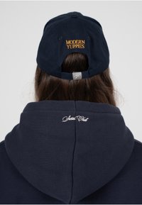 Marineblå baseballcaps med "MODERN YUPPIES" brodert i oransje, sammen med en marineblå hettegenser med "Social Club" sydd i hvitt.