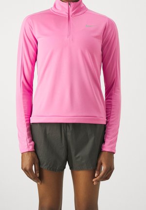 Persona que lleva una camiseta deportiva de manga larga de color rosa brillante con cremallera al cuarto y pantalones cortos de correr gris oscuro, de pie contra un fondo blanco.