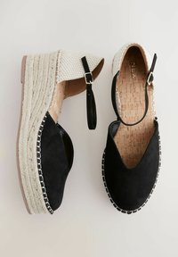 Espadrillas con tacco a zeppa in suede nero con dettagli in tessuto bianco intrecciato, cinturino alla caviglia, soletta in sughero e bordo intrecciato bianco e nero.