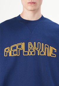 Voksen som har på seg en blå genser med rund hals og oransje omrissede tekst "REPLAY INC" over brystet.