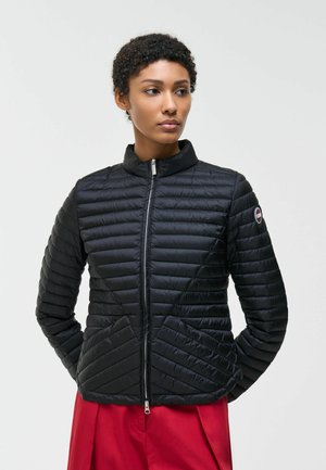 Donna con capelli corti e ricci, indossa una giacca nera trapuntata con zip e pantaloni rossi, guarda di lato con le mani nelle tasche.