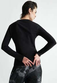 Top nero a maniche lunghe in tessuto morbido, design aderente, con scollatura tonda. La modella indossa pantaloni con paillettes, di texture, con un aspetto metallico.