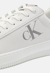 Baltas ādas sporta apavi ar teksturētu gumijas zoli, pelēku "ck" logo un plakanām baltām šņorēm. Iespēlē minimālistisku dizainu ar smalkiem akcentiem.