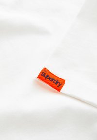 Esquina de una camiseta de algodón blanca con una etiqueta tejida naranja que dice "superdry" en azul, mostrando una textura suave y un diseño sencillo.