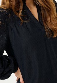 Blouse noire avec corps texturé et accents en dentelle sur les manches bouffantes, présentant un décolleté en V profond et une coupe décontractée.