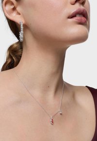 Collar de plata con un colgante de gema roja y una pequeña pieza de acento, usado junto a un aro de diamantes. Texturas suaves.