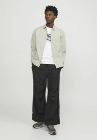 Chemise boutonnée gris clair portée sur un T-shirt graphique blanc, associée à un pantalon noir ample et des baskets grises. Design simple et coupe décontractée.