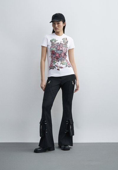 Νεαρή γυναίκα με λευκό μπλουζάκι με γραφικό σχέδιο σε κόκκινο και πράσινο, μαύρο παντελόνι cargo με φερμουάρ και αγκράφες, μαύρα παπούτσια και μαύρο καπέλο.