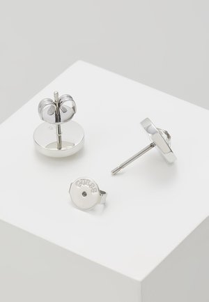 Boucles d’oreilles clous en métal couleur argent avec fermoir papillon et une pièce arrière gravée du mot « GUESS » sur une surface blanche.