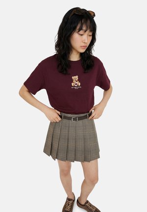 Jeune femme portant un t-shirt bordeaux avec un motif ours en peluche, une jupe plissée grise avec ceinture, des baskets marron et des lunettes de soleil sur la tête, mains sur les hanches.