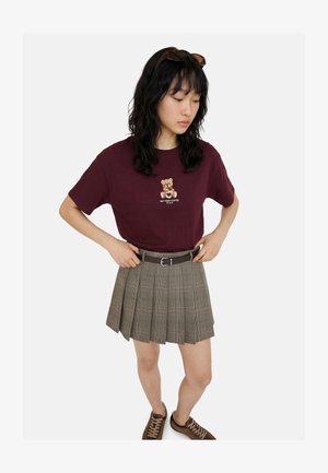 Jeune femme portant un t-shirt bordeaux avec un motif ours en peluche, une jupe plissée grise avec ceinture, des baskets marron et des lunettes de soleil sur la tête, mains sur les hanches.