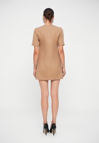 Figurpassad kort klänning i tan-färgad tyg med en mjuk textur. Har korta ärmar och rak fåll, matchad med svarta högklackade skor.
