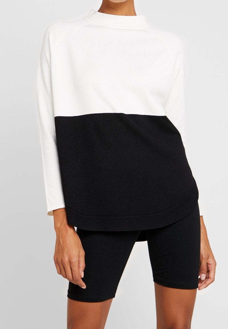 Personne portant un pull à manches longues blanc et noir en color block associé à un short noir ajusté sur un fond uni.