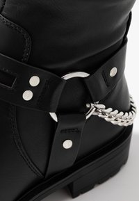 Stivale alla caviglia in pelle nera con rivetti argentati e dettagli decorativi in metallo a forma di anello e catena sul lato vicino al tallone.