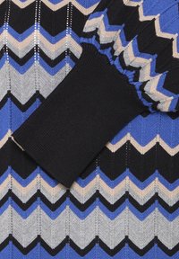Détail d'un pull en maille avec un motif chevron en bleu, gris et noir, finition texturée et un poignet côtelé noir.