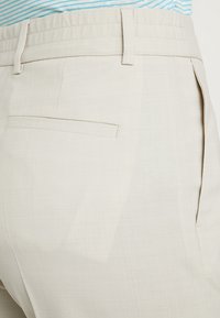 Beige broeken met een platte voorkant en achterzak, gemaakt van een textuurstof. De tailleband is elastisch voor comfort; zichtbaar zijn er horizontale strepen erboven.