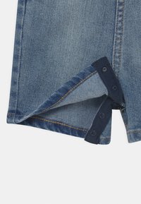 Džínová látka s vyšívanými oranžovými stehy a částečně rozepnutým tmavě modrým patentem na okraji oděvu.