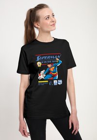 Černé bavlněné tričko s grafickým motivem Supermana s živými barvami, v komiksovém stylu, a textem "SUPERMAN NA NEBESÍCH" nahoře.