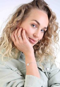 Zilveren armband met een klein bloemenbedel, gedragen om een pols. Krullend blond haar en een lichtgroene spijkerjasje zijn zichtbaar.