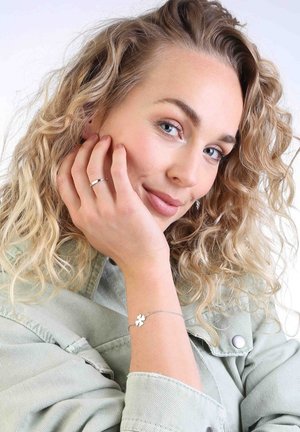 Bracelet en argent avec un petit pendentif en forme de fleur, porté au poignet. Des cheveux blonds bouclés et une veste en denim vert clair sont visibles.