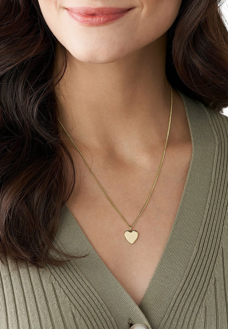 Femme portant un collier pendentif en forme de cœur en or avec une chaîne fine sur un pull vert côtelé à col en V.