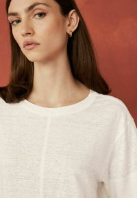 Tricou alb cu decolteu rotund și detalii vizibile de cusătură, realizat dintr-un material ușor. Modelul are părul castaniu de lungime medie.