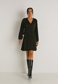 Πράσινο, μίνι φόρεμα με μακριά μανίκια, V-neck, κουμπιά στη λεπτομέρεια και ζώνη στη μέση. Συνδυάζεται με μαύρες μπότες μέχρι το γόνατο και μια μικρή τσάντα.