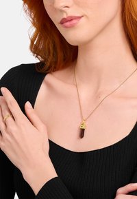 Goldener Anhänger mit braunem Kristall und sternförmiger Kappe, getragen über einem schwarzen gerippten Oberteil, kombiniert mit einem goldenen Ring.