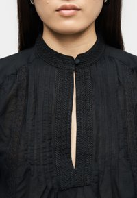 Κοντινό πλάνο ατόμου που φοράει μαύρο μπλουζάκι με λεπτομερή κέντημα, στρογγυλή λαιμόκοψη και μικρό κούμπωμα με κουμπί στο γιακά.