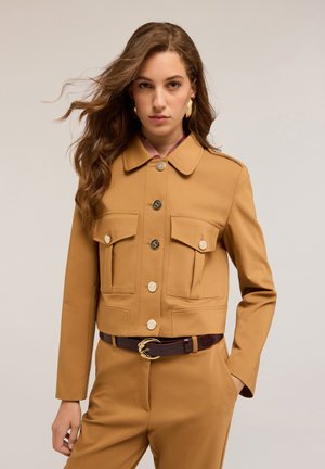 Gekürzter, brauner Blazer mit Kragen, zwei Brusttaschen und goldenen Knöpfen. Getragen über einer pinkfarbenen gemusterten Bluse und kombiniert mit einem dunklen Gürtel.