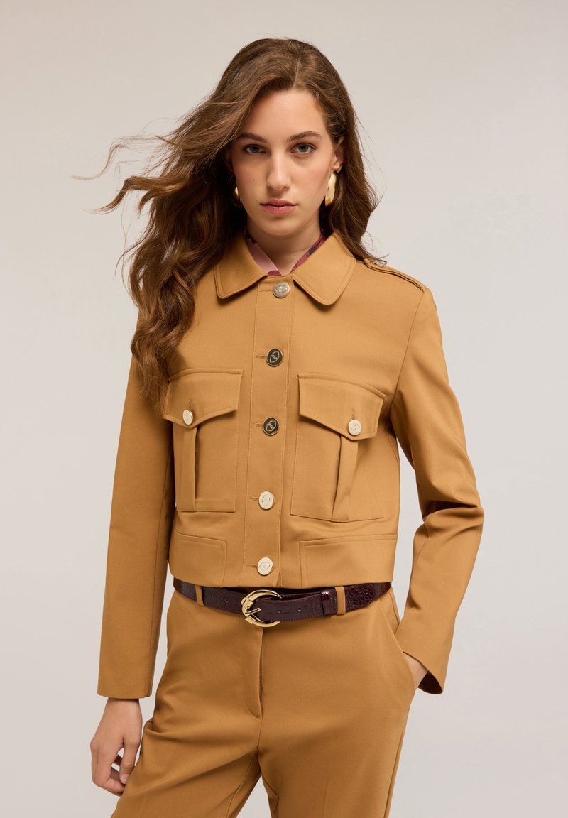 Giacca marrone tagliata con colletto, due tasche sul petto e bottoni dorati. Indossata sopra una blusa rosa con motivo e abbinata a una cintura scura.