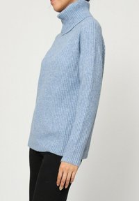 Femme portant un pull à col roulé côtelé bleu clair et un pantalon noir, vue de côté sur un fond uni.