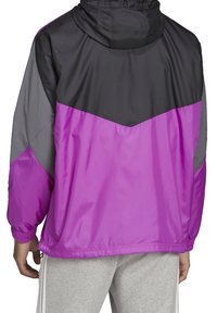 Chaqueta cortavientos con bloques de color que presenta un diseño en negro, gris y un vibrante morado, con capucha y puños elásticos. Material ligero.