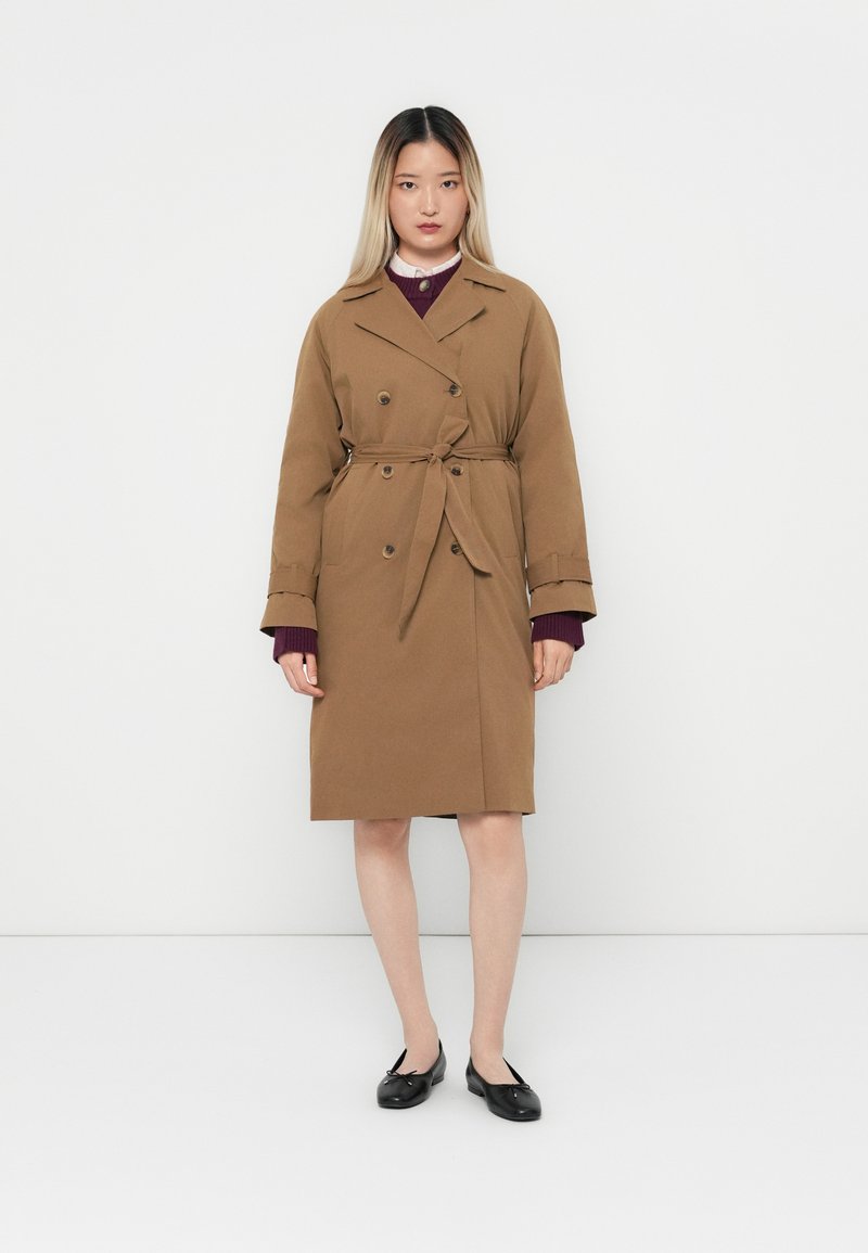 Brun trenchcoat med bälte i midjan, dubbelknäppt design och justerbara ärmar. Bärs över en börrgundy-plagg, i kombination med svarta skor.