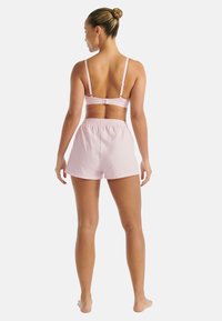 Kobieta stojąca boso tyłem, ubrana w jasnoróżowy stanik sportowy i luźne szorty na tle jednolitej białej ściany.