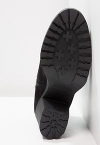 Svart mockaskor med en texturerad gummisålemönster i plustecken. Klacken är platt och med varumärket synligt på sålen.