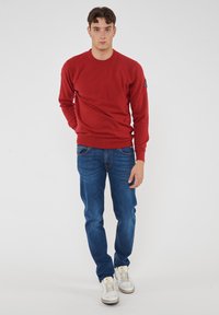 Maglione rosso a girocollo con polsini e orlo a costine, abbinato a jeans in denim blu e sneakers bianche. Il design è semplice e aderente.