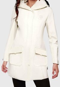Kobieta w kremowym płaszczu z kapturem, zapinanym na zatrzaski z przodu i dwoma kieszeniami z patkami, w połączeniu z czarnymi spodniami.