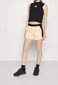 Mujer vistiendo un top corto sin mangas negro y shorts deportivos durazno con lados negros, combinados con calcetas negras y zapatillas negras.