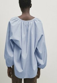 Personne portant une blouse ample à manches longues bleu clair avec un col froncé, vue de dos sur un fond uni.