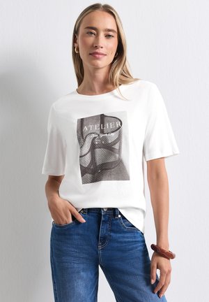 Kvinde med glat blondt hår iført hvid kortærmet skjorte med grafisk tryk og blå jeans, stående med den ene hånd i lommen.