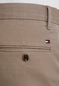 Písečné látkové kalhoty s hladkým povrchem, mají zadní kapsu na knoflíkové zapínání a malé vyšívané logo v červené, bílé a modré barvě.