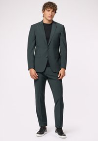 Completo verde scuro realizzato in tessuto liscio, caratterizzato da un blazer aderente con un singolo bottone e pantaloni abbinati, abbinato a sneakers nere.