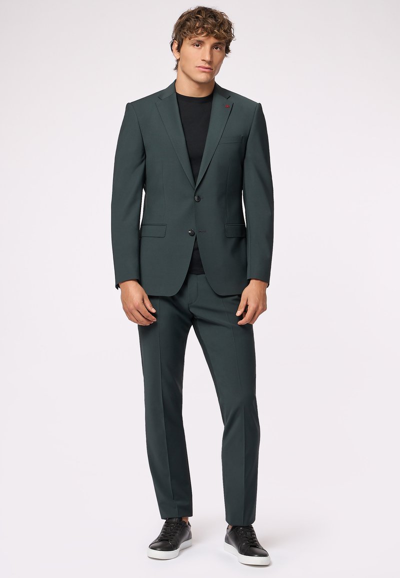 Completo verde scuro realizzato in tessuto liscio, caratterizzato da un blazer aderente con un singolo bottone e pantaloni abbinati, abbinato a sneakers nere.